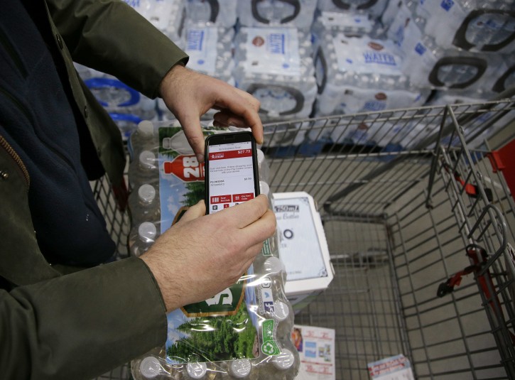 In this Tuesday, Feb. 13, 2018 photo Tony D'Angelo uses the BJ's Express Scan app on his cell phone to scan a case of bottled water he is purchasing while shopping at the BJ's Wholesale Club in Northborough, Mass.  Many stores are letting customers scan their shopping choices with an app on their phone or the store's mobile device as they roam in and out of the aisles before finalizing their purchases at an express checkout. (AP Photo/Stephan Savoia)