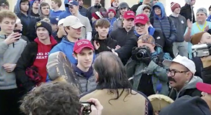 In this Friday, Jan. 18, 2019 image made from video provided by the Survival Media Agency, a teenager wearing a "Make America Great Again" hat, center left, stands in front of an elderly Native American singing and playing a drum in Washington. AP